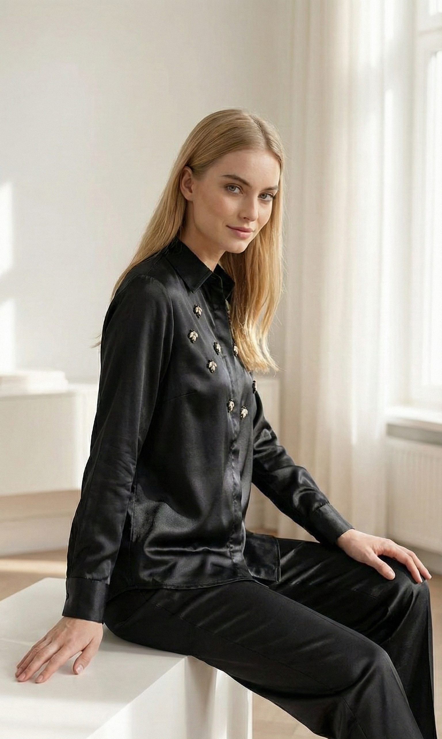 Blonde woman in black satin blouse with embellishments and matching black pants sitting indoors