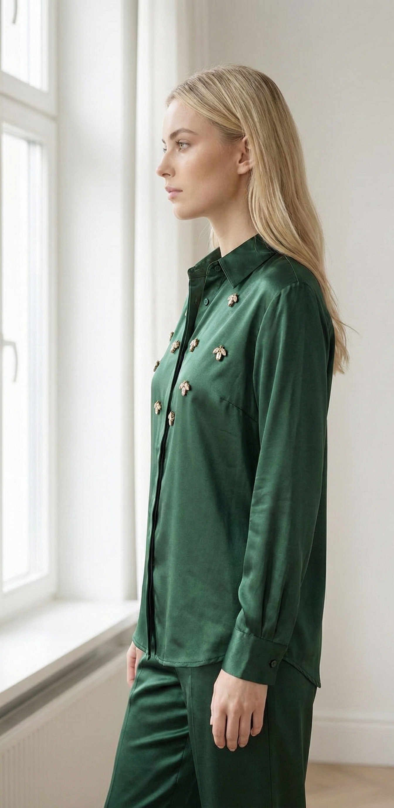 Blonde woman in green satin blouse and pants with bee embellishments standing by bright window