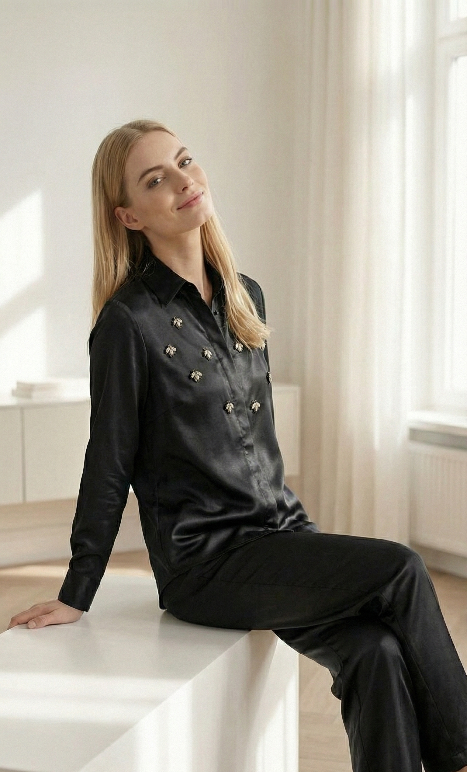 Blonde woman in black satin blouse with bee embroidery and black pants sitting in bright minimalist room